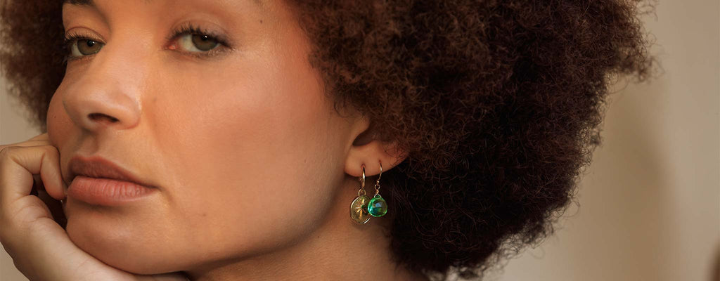 An ear stack featuring the Terra Drop Hoop Earrings, a gold disc features a palm tree, dangling from a golden hoop. These are styled beside green quartz gemstone drop earrings.