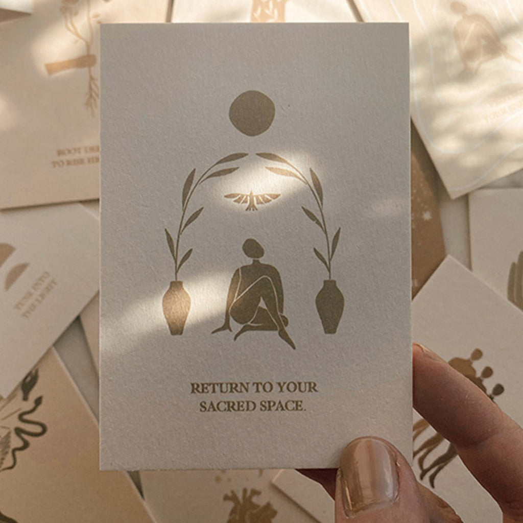 a card featuring a person surrounded by nature, ready for meditation. The affirmation on the card reads 'return to your sacred space'. This is one of the affirmations included in this deck of cards for practicing mindfulness and manifestation.