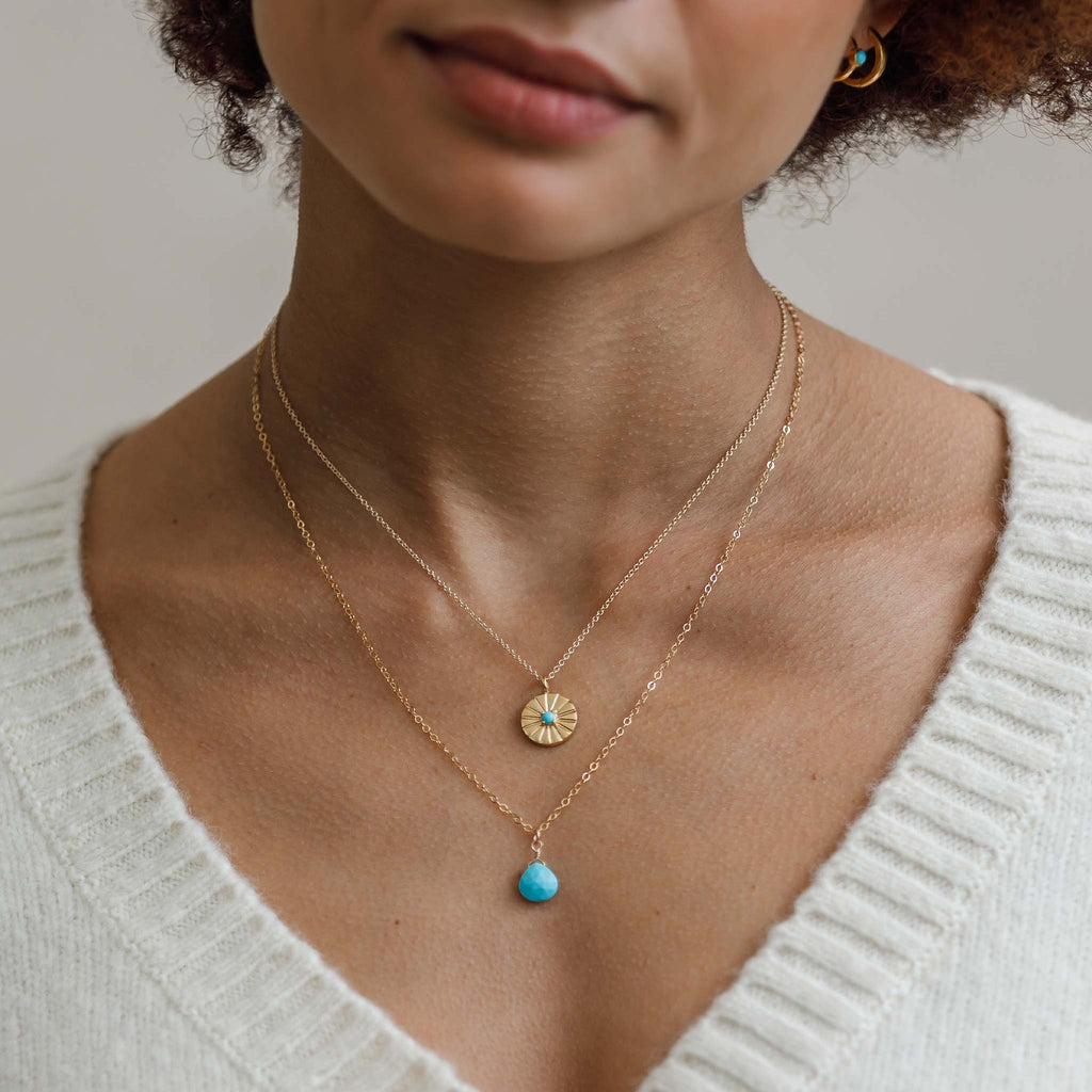 An Arizona Turquoise blue gemstone hands from a minimal gold chain. This gemstone pendant necklace is a colourful and modern necklace, styled with the December Turquoise Mini Sundial Necklace in a gold toned necklace stack. Shop meaningful gemstone jewellery.