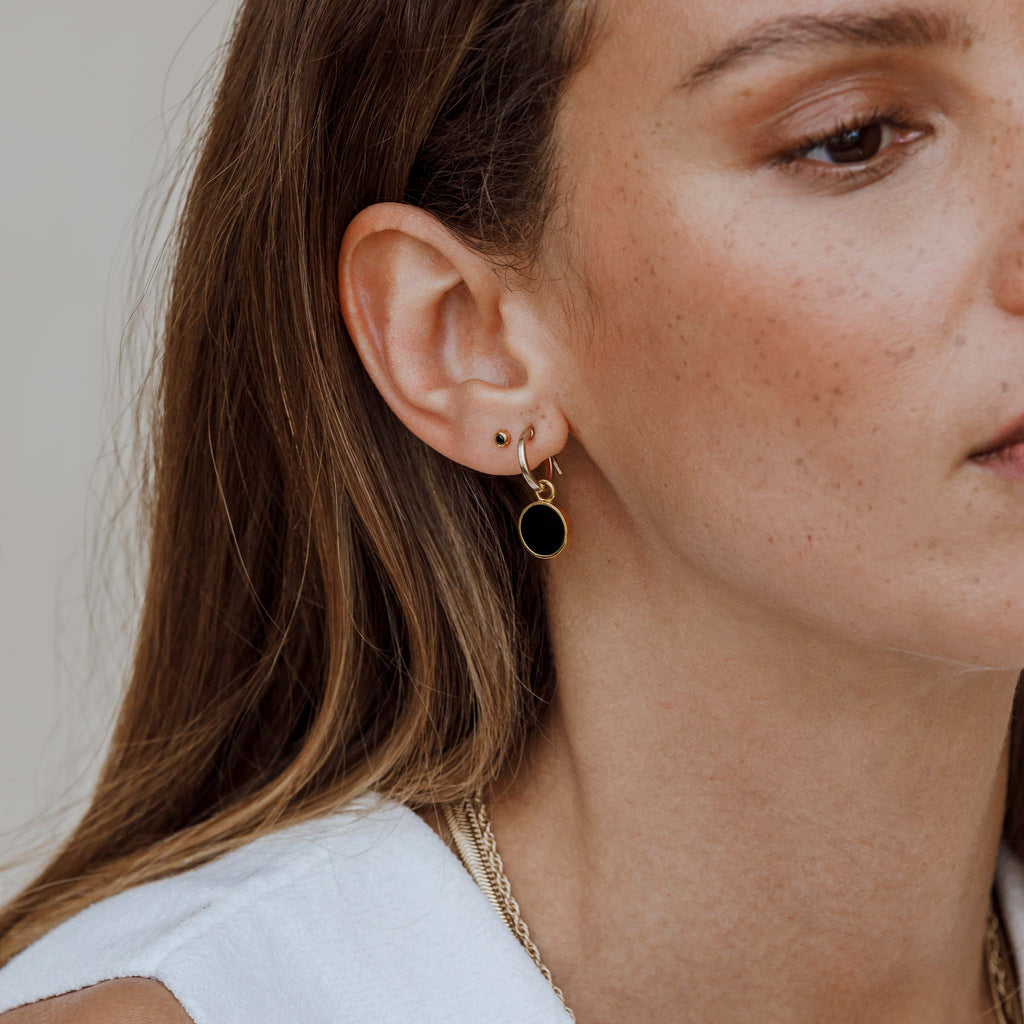 Slices of black onyx gemstones are set in a gold circular frame. These gemstone charms hand from gold creole hoop earrings. Styled into a matching ear stack, the Portilla hoop earrings are worn beside the Black Onyx Punta Stud Earrings. Shop 14k gold vermeil, gemstone jewellery designed in the UK.