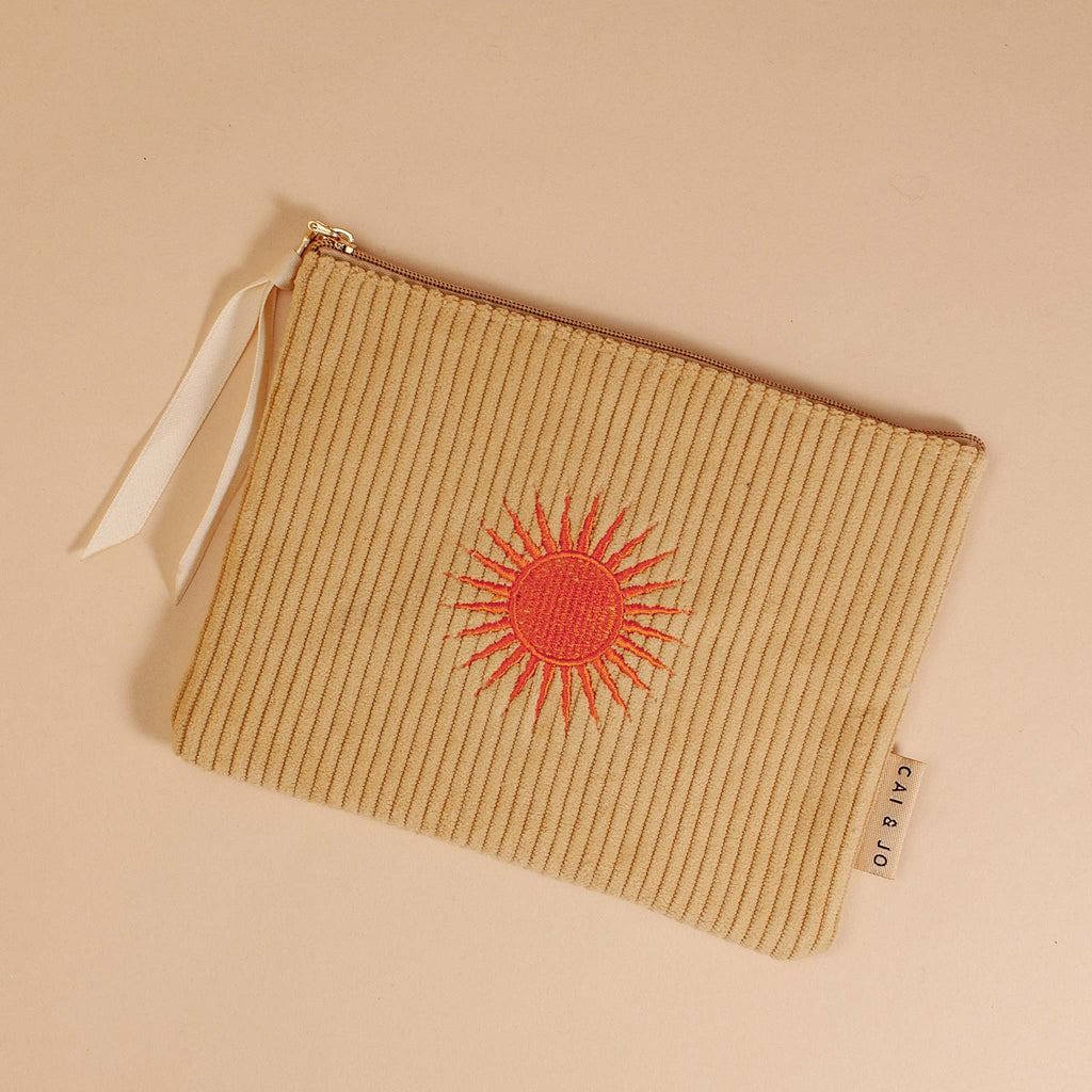 A flat corduroy pouch in a pale ochre colour, featuring an embroidered orange sun motif.