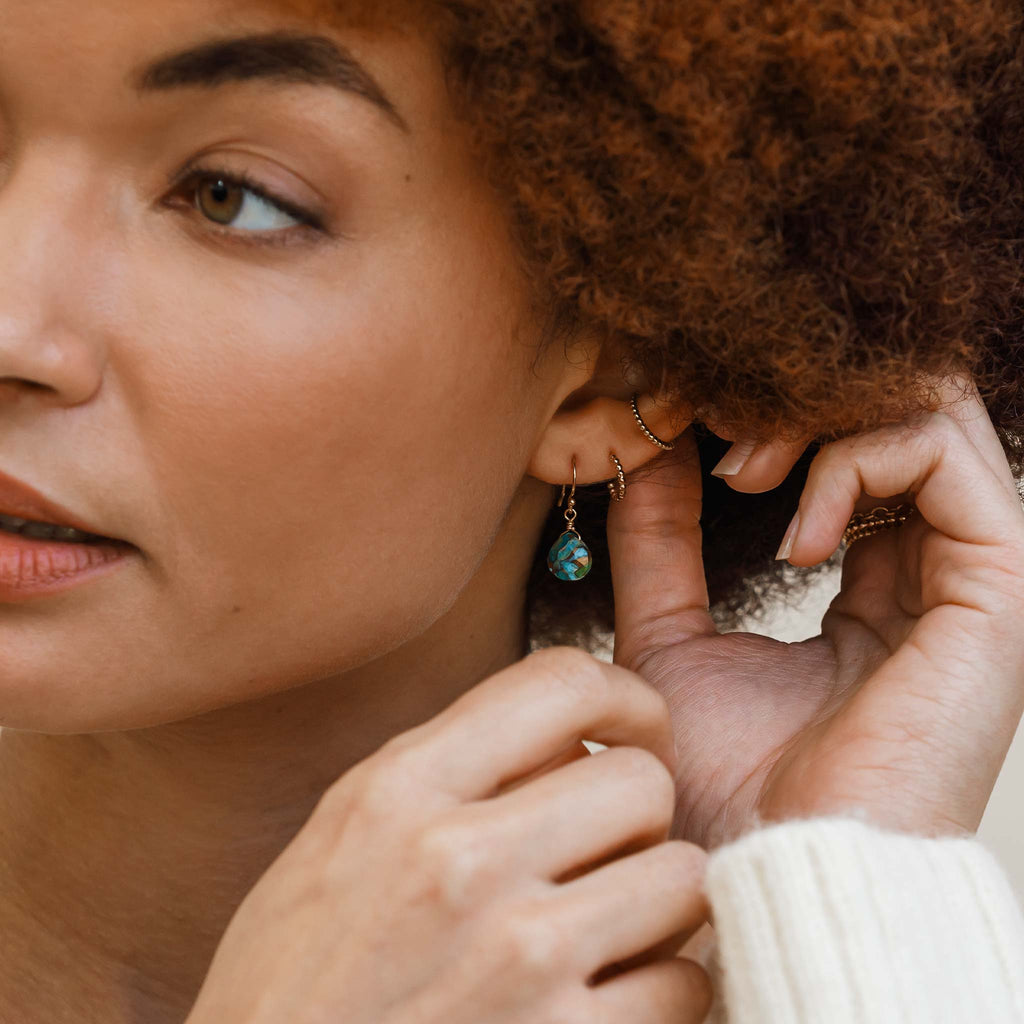 Model wears the Isla Drop gemstone earrings with faceted mohave gemstones dropping from 14k gold fill earwire.This gemstone features organic patterns of gold running throughout each facet. Styled with minimal hoops.