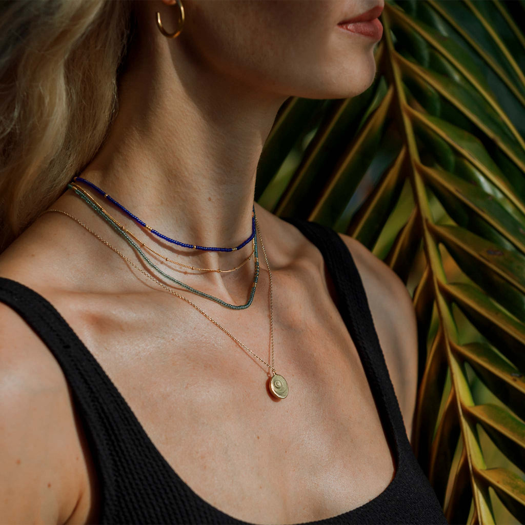 The Morocco Blue Beaded Necklace features bright cobalt blue beads, contrasted with flecks of gold beads. This choker length necklace is seen styled with another beaded necklace, a gold layering chain and a gold shell pendant necklace.
