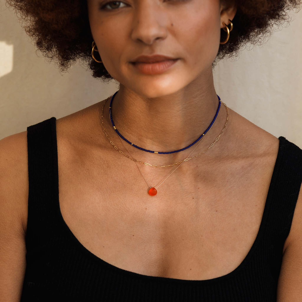 A sunset orange carnelian gemstone floats on a fine cord, forming and minimal and modern gemstone necklace. Styled with a gold paperclip style layering chain and a dark blue beaded necklace. 