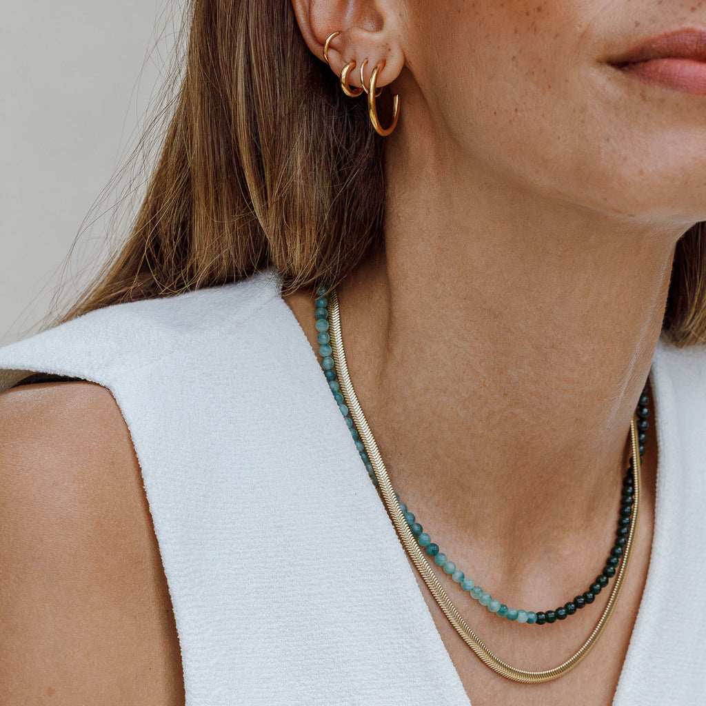 The Paseo Herringbone Chain Necklace is a wide snake chain necklace, with a metallic and light-reflecting finish. This iconic and timeless chain can be worn solo or in a necklace stack. Worn here with the Accoya Jade Beaded Gemstone Necklace.