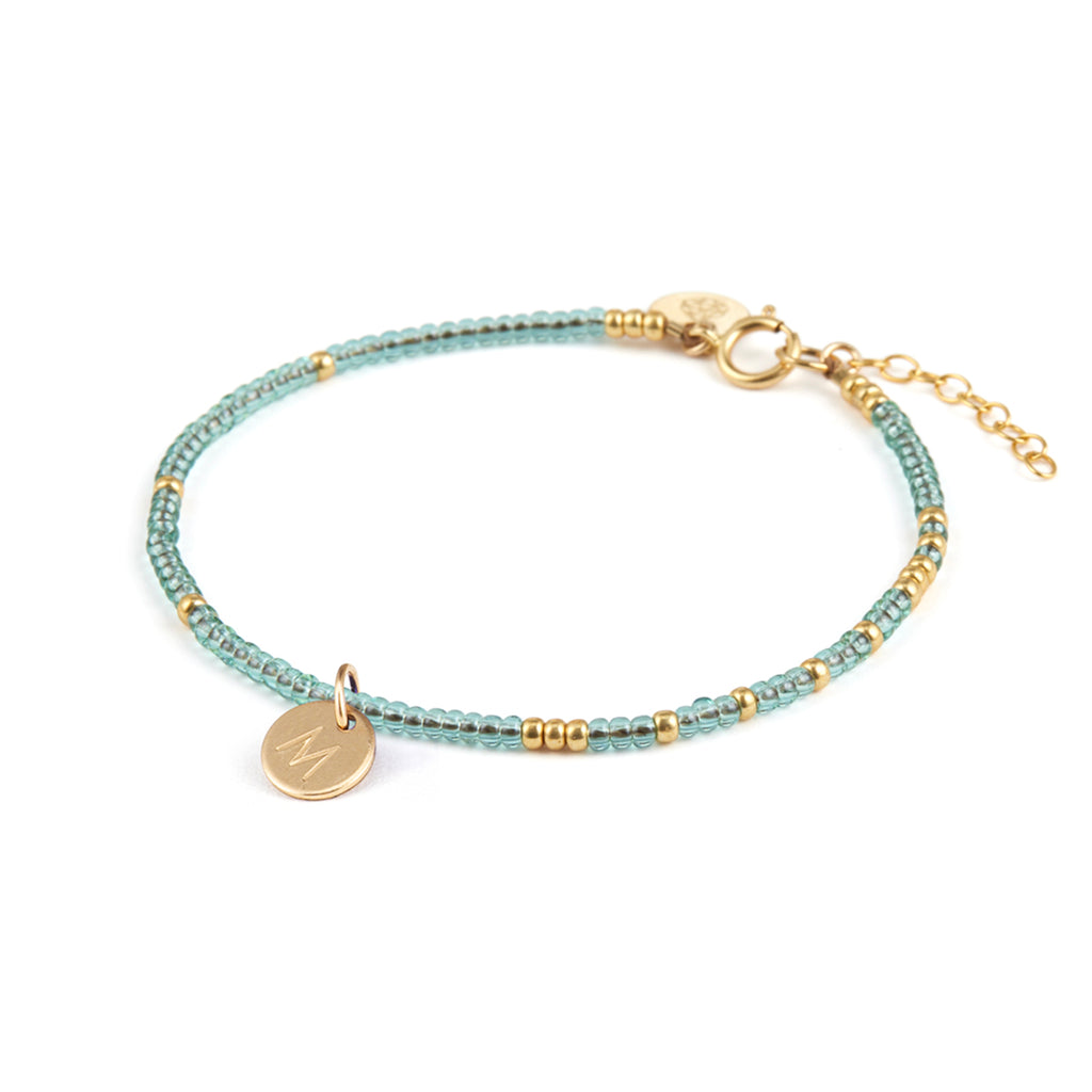 Personalised with a round charm, hand-stamped with an M, the water blue bracelet is made from blue and gold seed beads. 