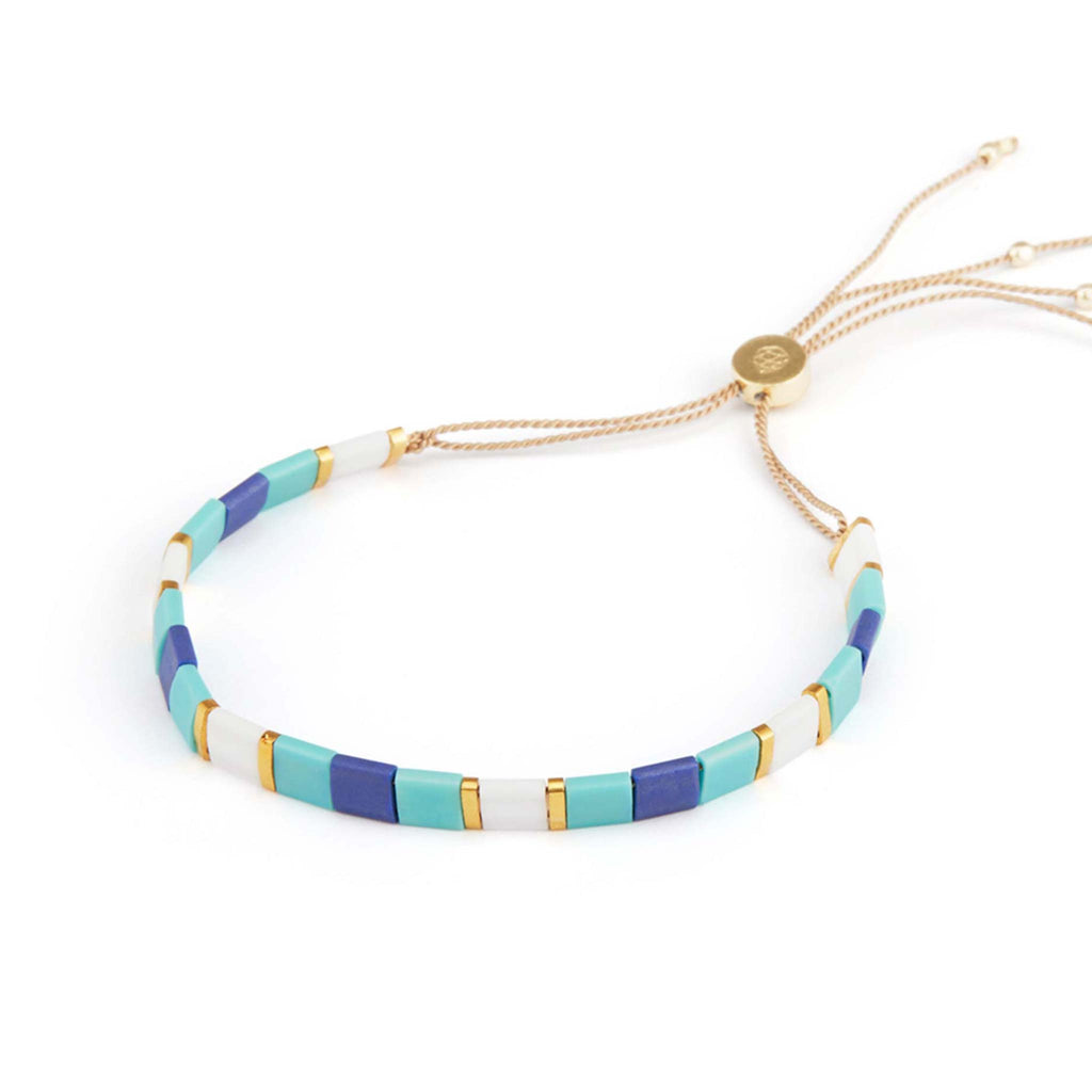 Alternating glass Tila beads in turquoise, cobalt blue, gold and white create a pattern in this beaded bracelet. Featuring a slider closure on silk, this bracelet is adjustable and adds an effortless pop of colour summer.