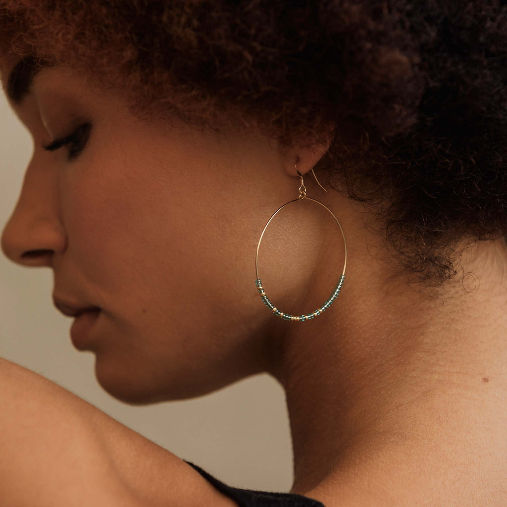 Model wears large beaded hoop earrings. The Lido Hoops feature translucent beads in water blue, contrasted with gold beads.