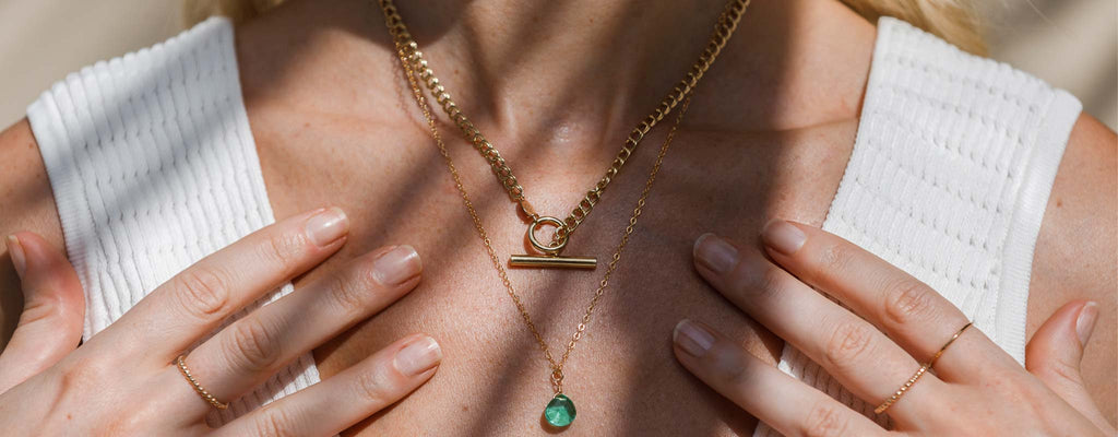Model wears a layered necklace stack with a gold vermeil double curb necklace featuring a t-bar toggle fastening, and a green quartz gemstone necklace.
