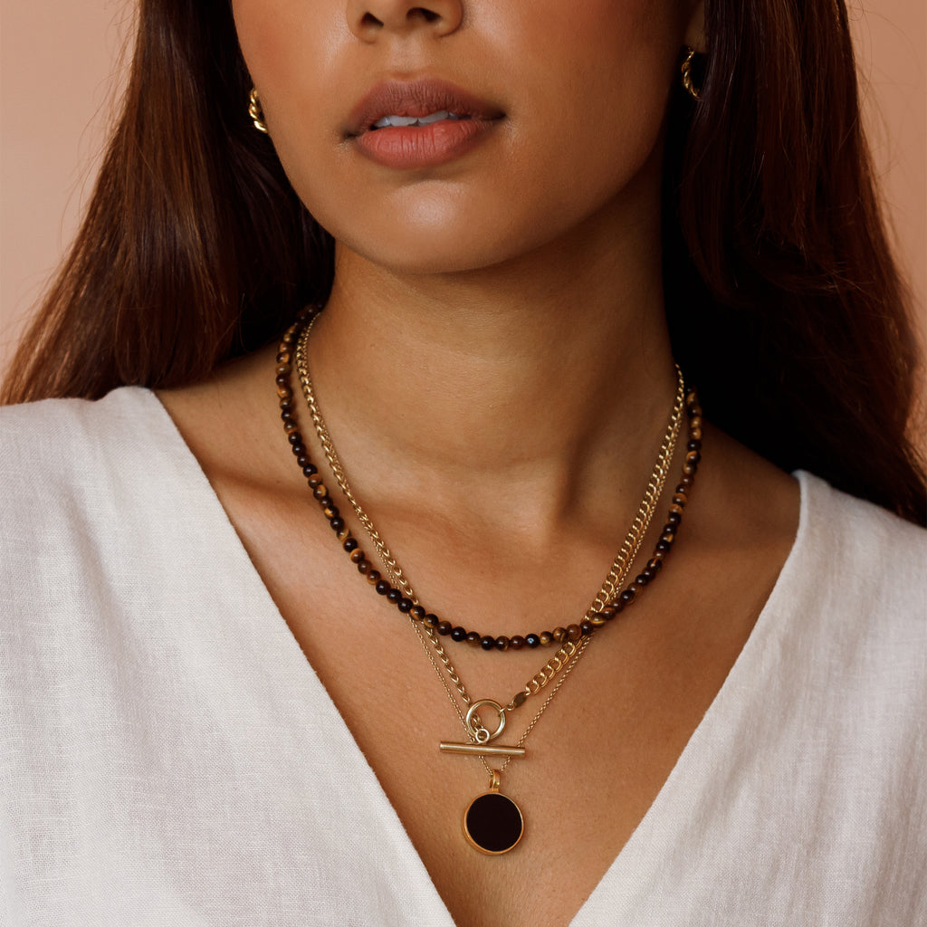 A layered necklace stack featuring Tiger’s Eye Āina Beaded Necklace, Morgan Double Curb Toggle Necklace and the Black Onyx Porthole Necklace.
