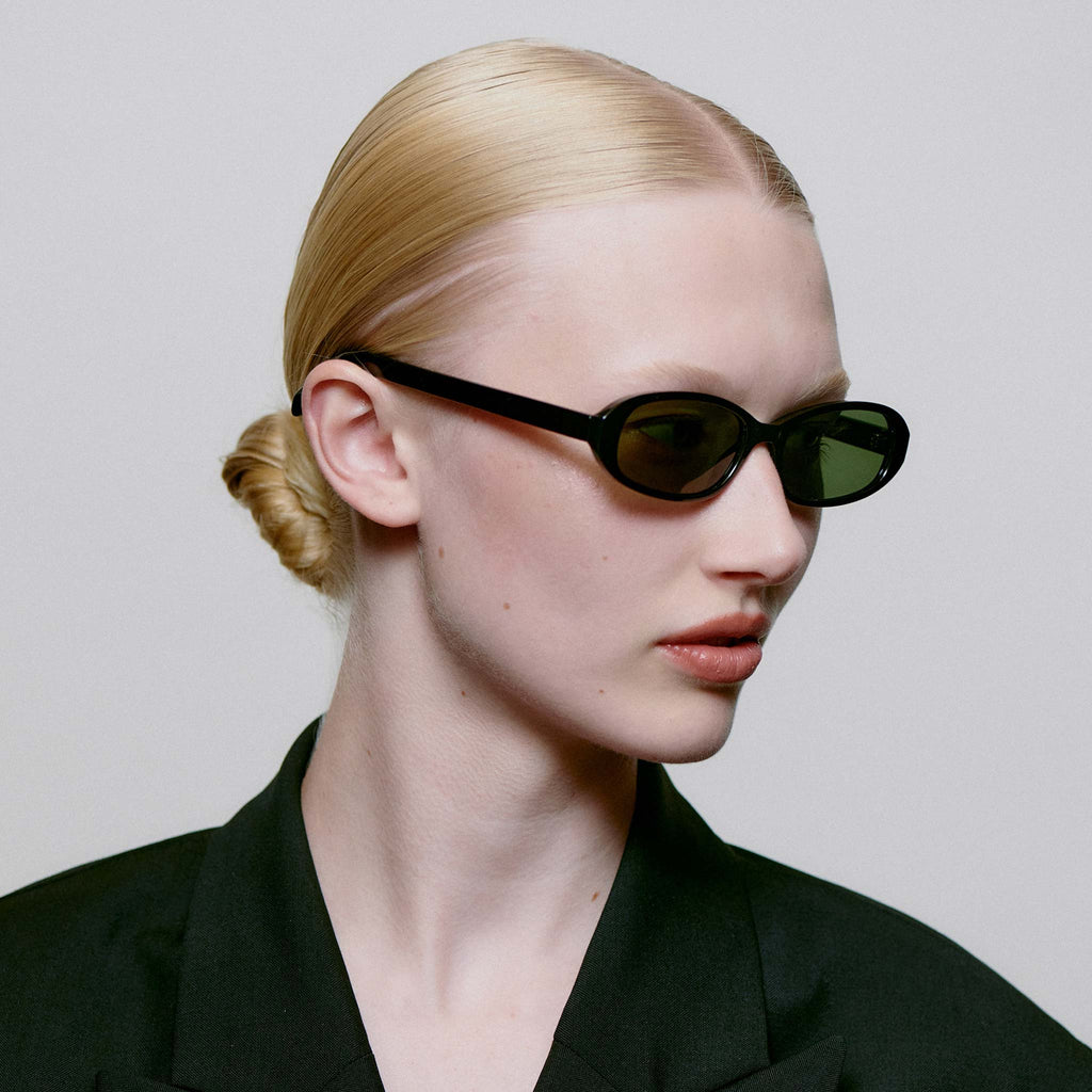 Person wearing black sunglasses and a black outfit against a plain background.