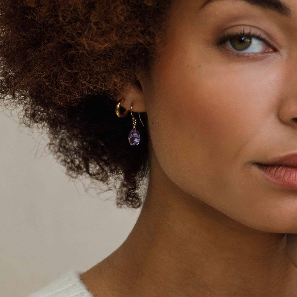 An Alexandrite Quartz Gemstone dangles from an ear wire. Alexandrite Quartz is a gemstone that appears both blue and purple hues in different lights. These modern and minimal drop earrings are styled in an ear stack with a minimal hoop earring.