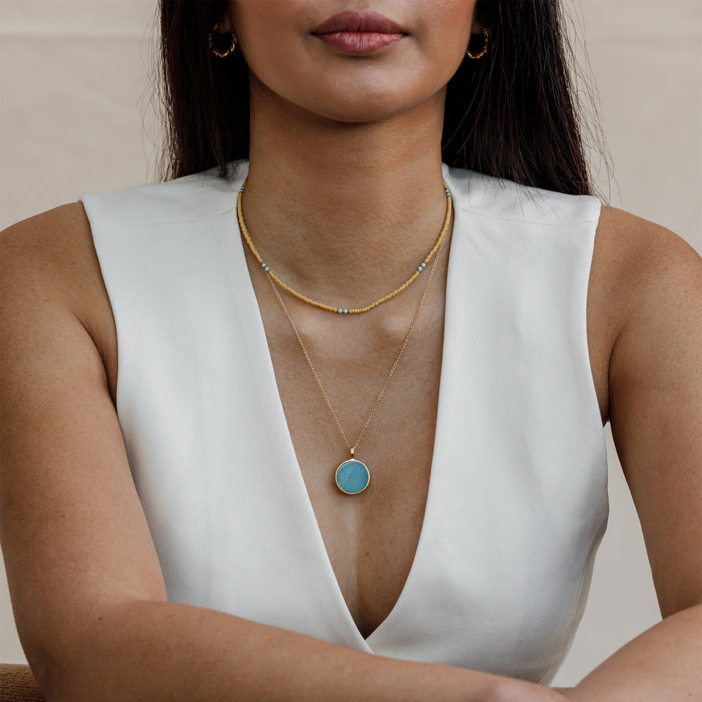 A slice of Amazonite is set in a circular brushed gold vermeil pendant. The turquoise toned pendant hangs on a minimal gold fill chain, styled with a yellow beaded choker necklace.