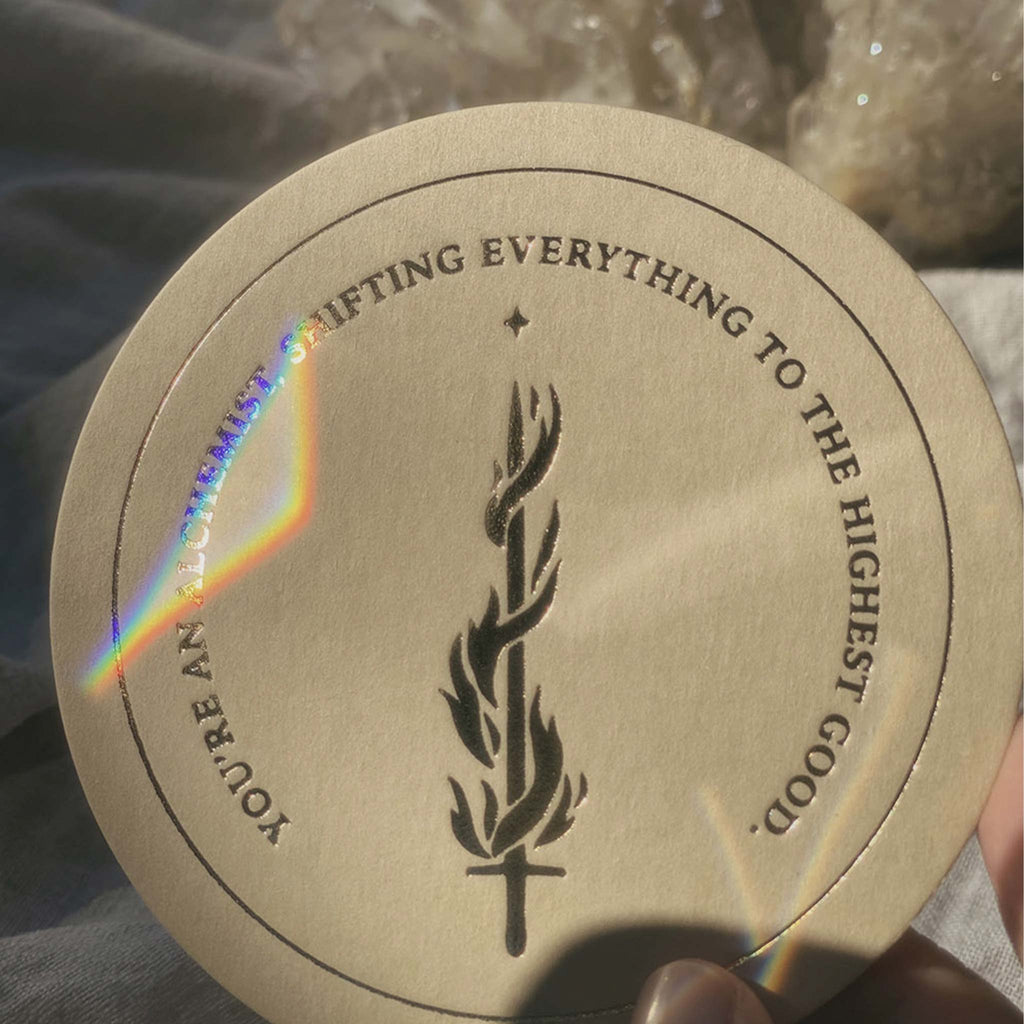 'You're an alchemist, shifting everything to the highest good' is printed in gold-foil press on a circular card alongside an illustration of a sword on fire with a spark.