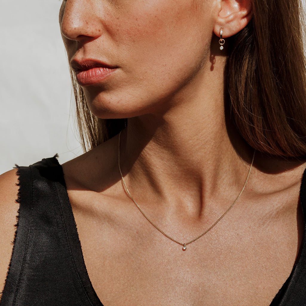 Model wears Axios diamond huggie hoop earrings and then coordinating diamond necklace on a flat curb chain. 