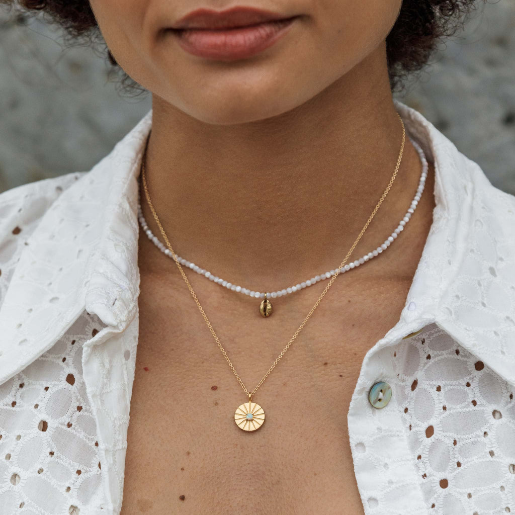 A dainty hand-cast cowry shell dances on a mother of pearl beaded choker necklace. Styled in a layered necklace look with a gold pendant necklace.