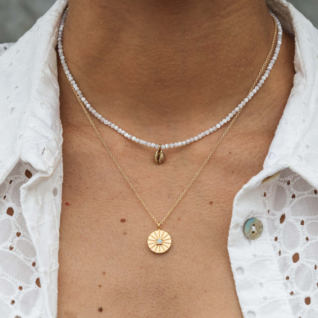 A dainty hand-cast cowry shell dances on a mother of pearl beaded choker necklace. Styled in a layered necklace look with a gold pendant necklace.