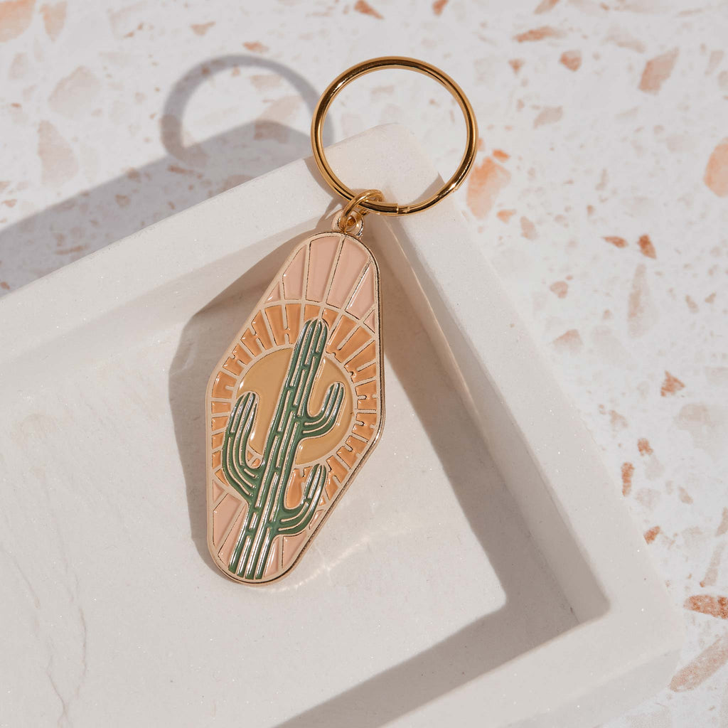 A gold plated motel key style key chain features a cactus and sun-ray design, made with soft enamel and gold plating.