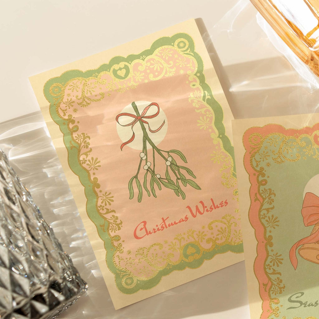 A Christmas greetings card reading 'Christmas Wishes', featuring festive mistletoe, accented by green, pink and gold foil. Designed by Cai and Jo, available at Wanderlust Life.