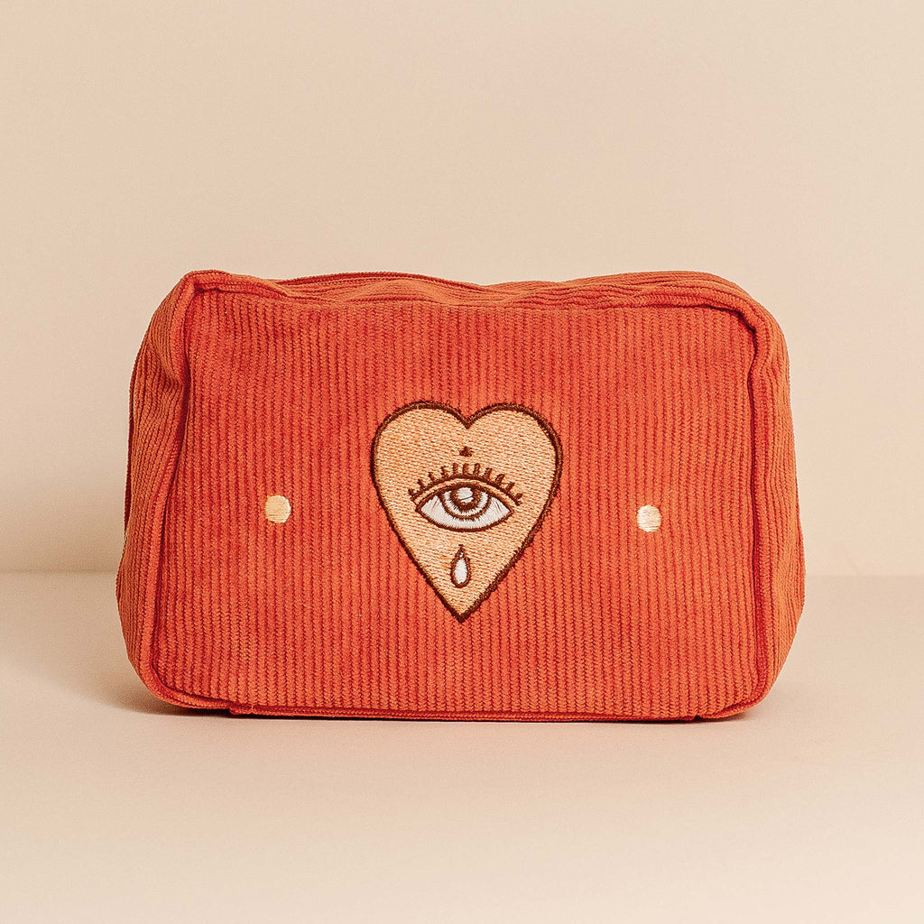 A rusty pink and red toned makeup bag, made with textured corduroy fabric. Featuring a zip closure and internal pockets, perfect for organising cosmetic products on the go. This makeup bag features an embroidered motif with an eye surrounded by a heart.