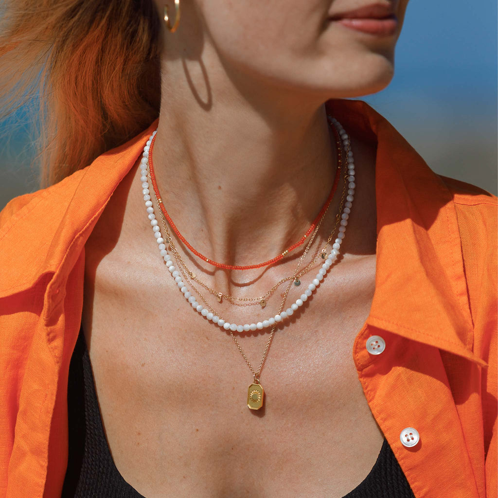 The Kai necklace features mother of pearl gemstone beads. Styled with an orange shirt and in a layered necklace stack with an orange beaded necklace and a golden charm necklace and pendant.