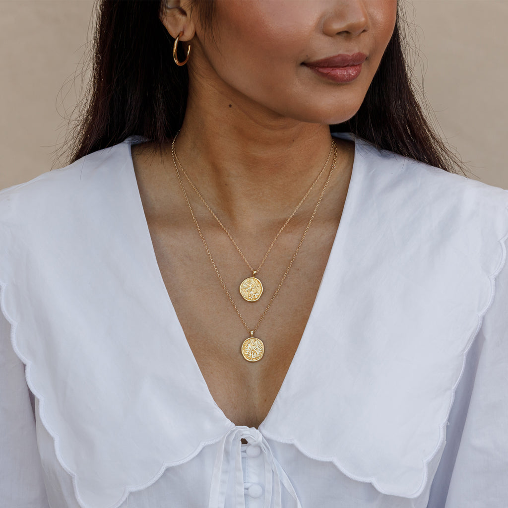 A layered necklace stack style two zodiac pendant necklaces layered together. Shaped like molten coins, the pendants are embossed with designs representing Sagittarius and Libra Zodiac signs.