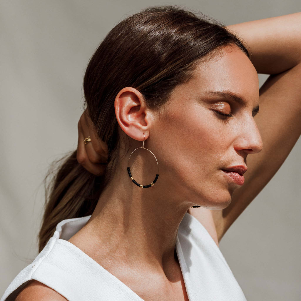 The Lido Noir Hoop Earrings feature black and gold beads, creating contrast between golden tones and darkness. These large hoops are a bold statement earring that can be worn as your daily staples or styled into an evening look.