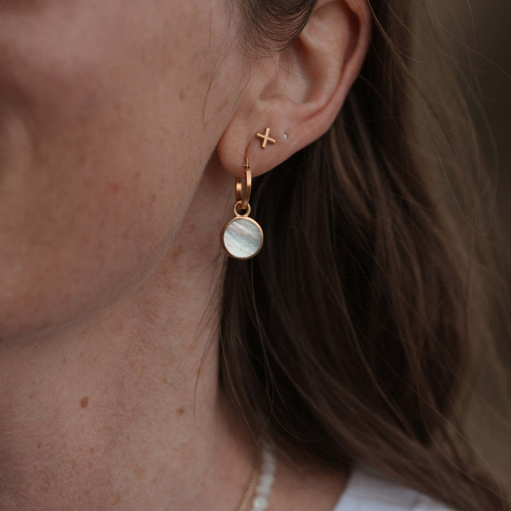 Iridescent Mother of Pearl is surrounded by a circular frame of brushed gold, reminiscent of light through a porthole. These charms hang from minimal gold vermeil hoop earrings. Styled in an ear stack with x studs.