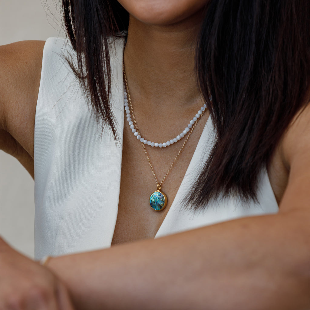 A Porthole Necklace with a circular slice of Paua shell, framed in brushed gold vermeil pendant. Styled with a mother of pearl beaded necklace.