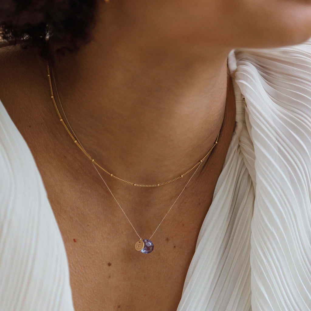 The Alexandrite Quartz Fine Cord Necklace features a blue and purple toned gemstone, strung on a minimal fine cord, creating the illusion of a floating gemstone. This minimal necklace is personalised with an initial tag, styled with a gold layering chain.