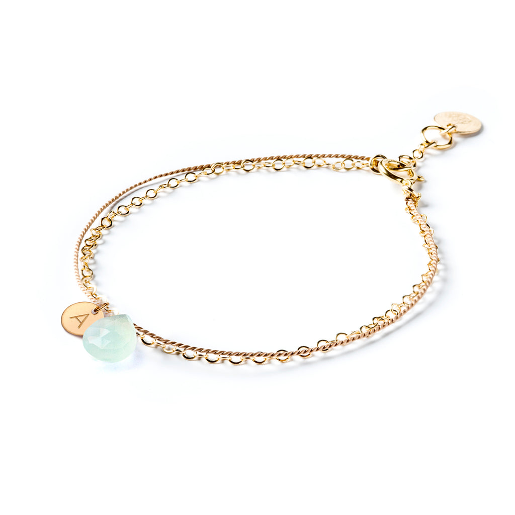 Personalised Sea Glass Chalcedony Gemstone Bracelet. A monogram tag is seen beside a sea foam blue gemstone, strung on silk and layered with a fine gold chain. Shop meaningful and personalised gemstone jewellery online at Wanderlust Life.