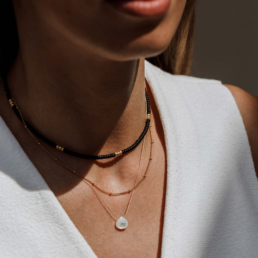 The Poolside Noir is a beaded necklace featuring small black beads, accented with gold beads creating a contrasting pattern. Styled in a necklace stack with the Satellite layering chain and mother of pearl fine cord necklace.
