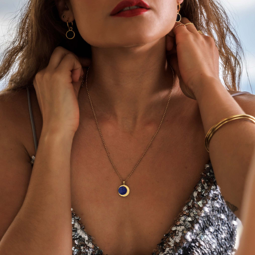 A circular slice of sodalite sits within a delicate round pendant surrounded by tiny rays etched into its surface forming a crescent silhouette that captures and reflects light with every movement. 