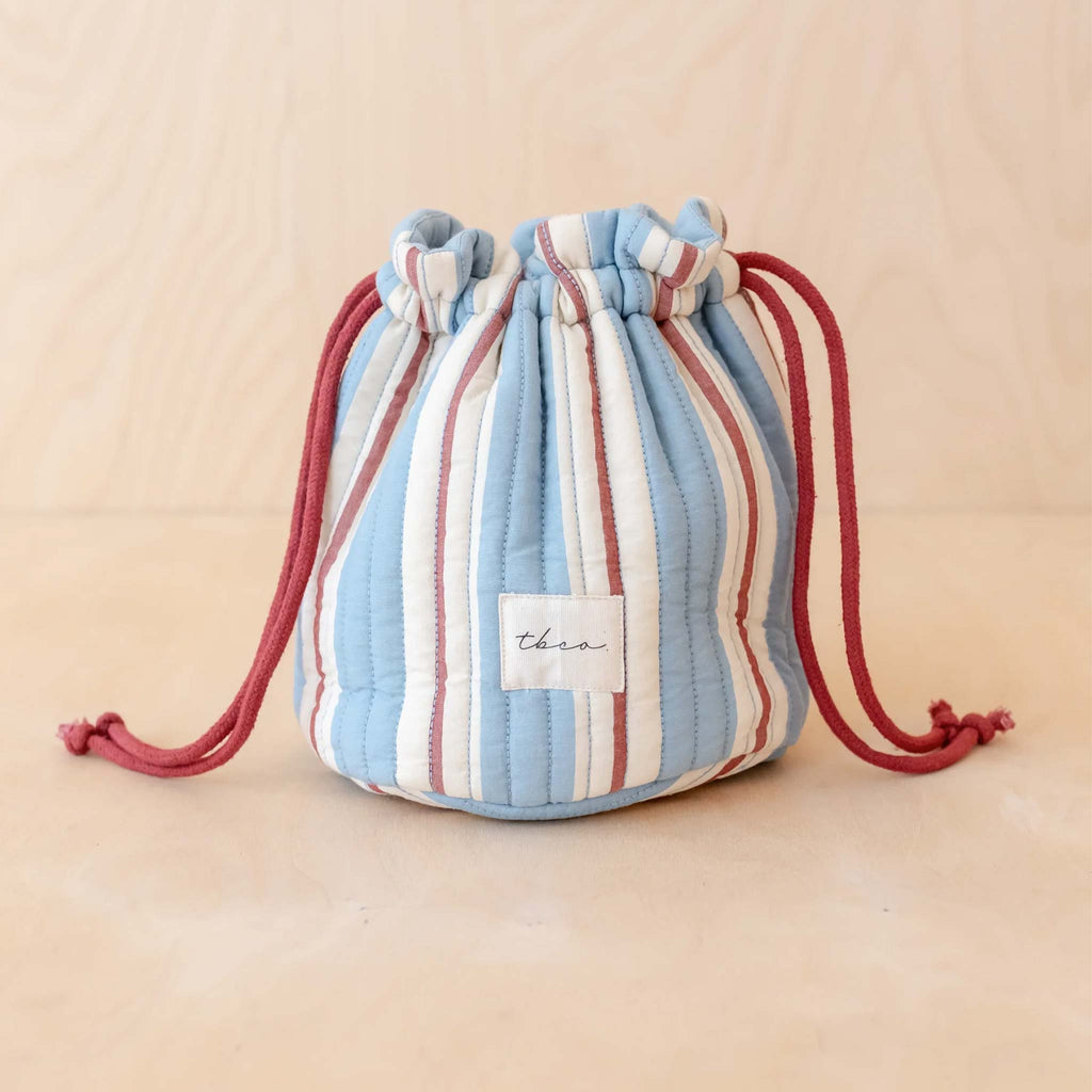 A quilted wash bag made with 100% cotton, featuring a red, blue and cream striped pattern, an embroidered TBCO logo and a draw string closure.