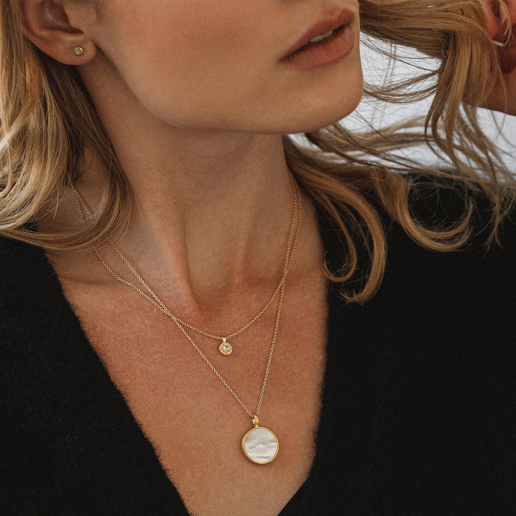 Mother of Pearl gemstone porthole necklace. A circular pendant with a mother of pearl slice is framed by gold with a brushed finish; this statement pendant necklace is on a long minimal chain. Wear solo or style with shorter necklaces to create a layered necklace stack with more impact.