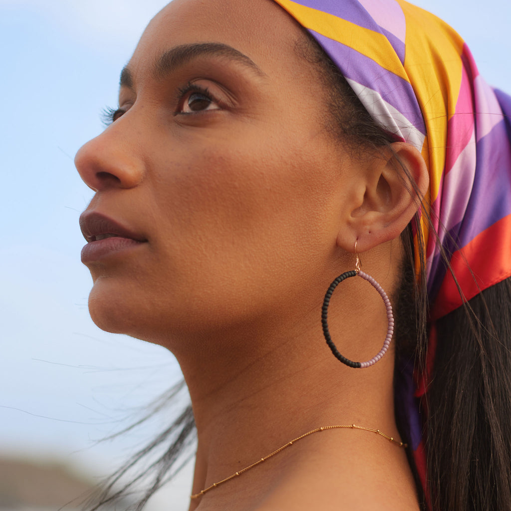 Model wears Lilac Lido Earrings. Black and lilac beads make a monochrome contrast in these statement large hoops. A summer must-have for beaded jewellery. Designed to effortlessly wear your jewellery from day to night, these statement hoops add effortless style to your summer style.
